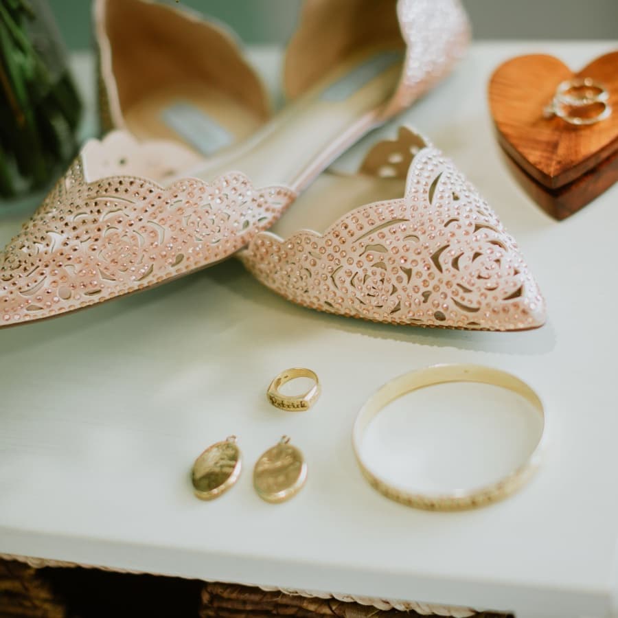 A pair of intricately designed pink shoes beside gold jewelry including a ring, earrings, and a bracelet on a white surface.