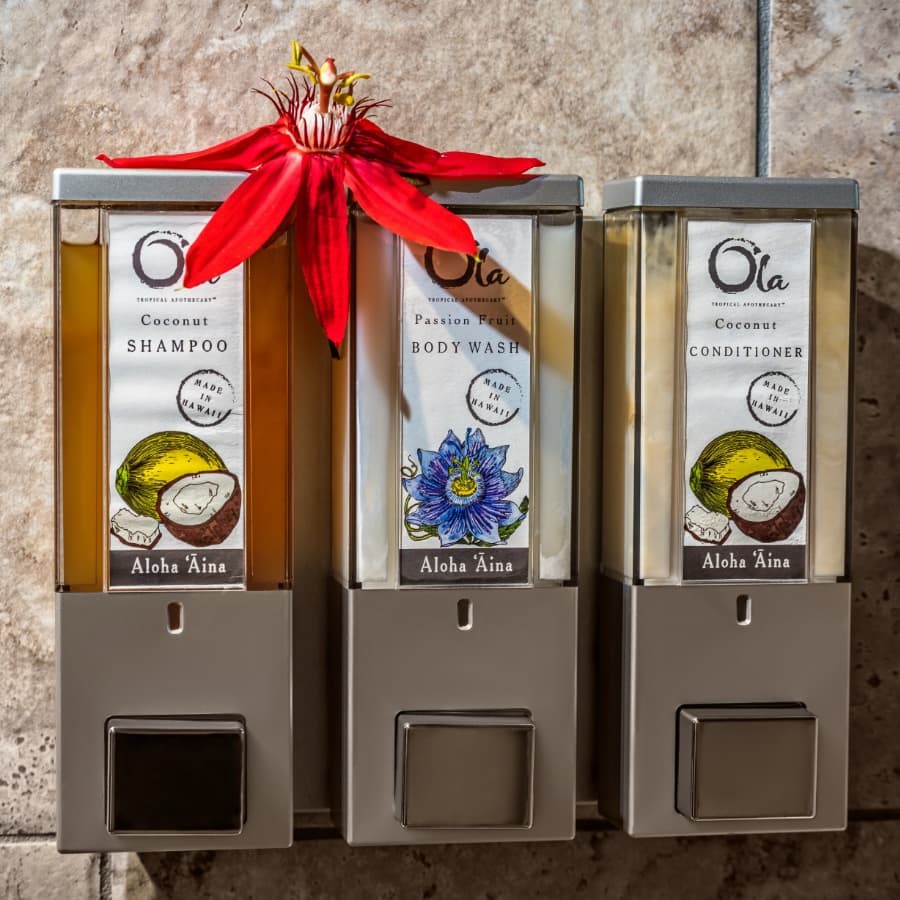 Three refillable shower dispensers filled with coconut shampoo, passion fruit body wash, and coconut conditioner, topped with a red flower.