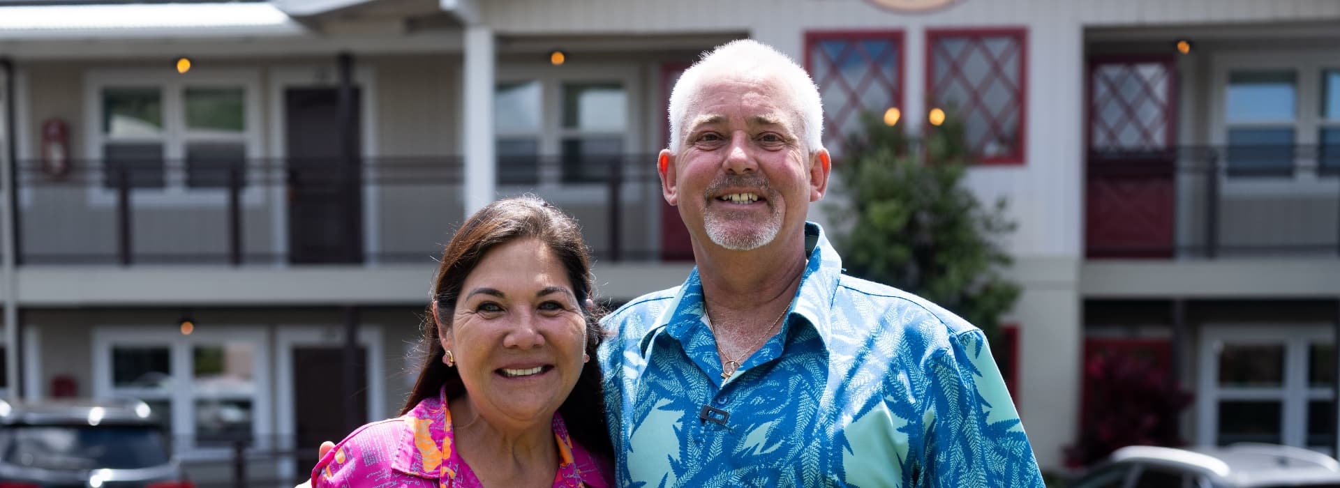 Two smiling individuals pose together outdoors in colorful tropical shirts, with a building in the background.