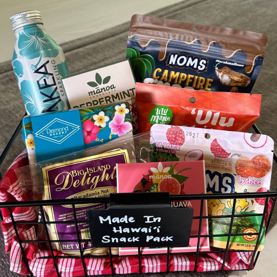 A wire basket filled with various snacks and treats from Hawaii, including macadamia nuts, guava, and a sparkling water.