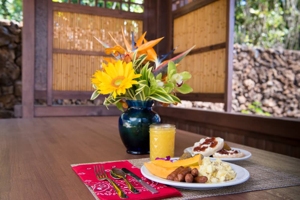 A beautifully arranged breakfast with tropical fruit, eggs, and a drink, complemented by a vibrant flower vase, sits on a wooden table.